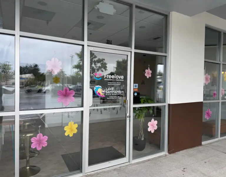 The storefront of Lake Sammamish Kids Dentistry - Pediatric Dentist in Sammamish, WA features large glass windows and a door adorned with colorful flower decals. Signs on the door include "Freelove" and the dental office's name.