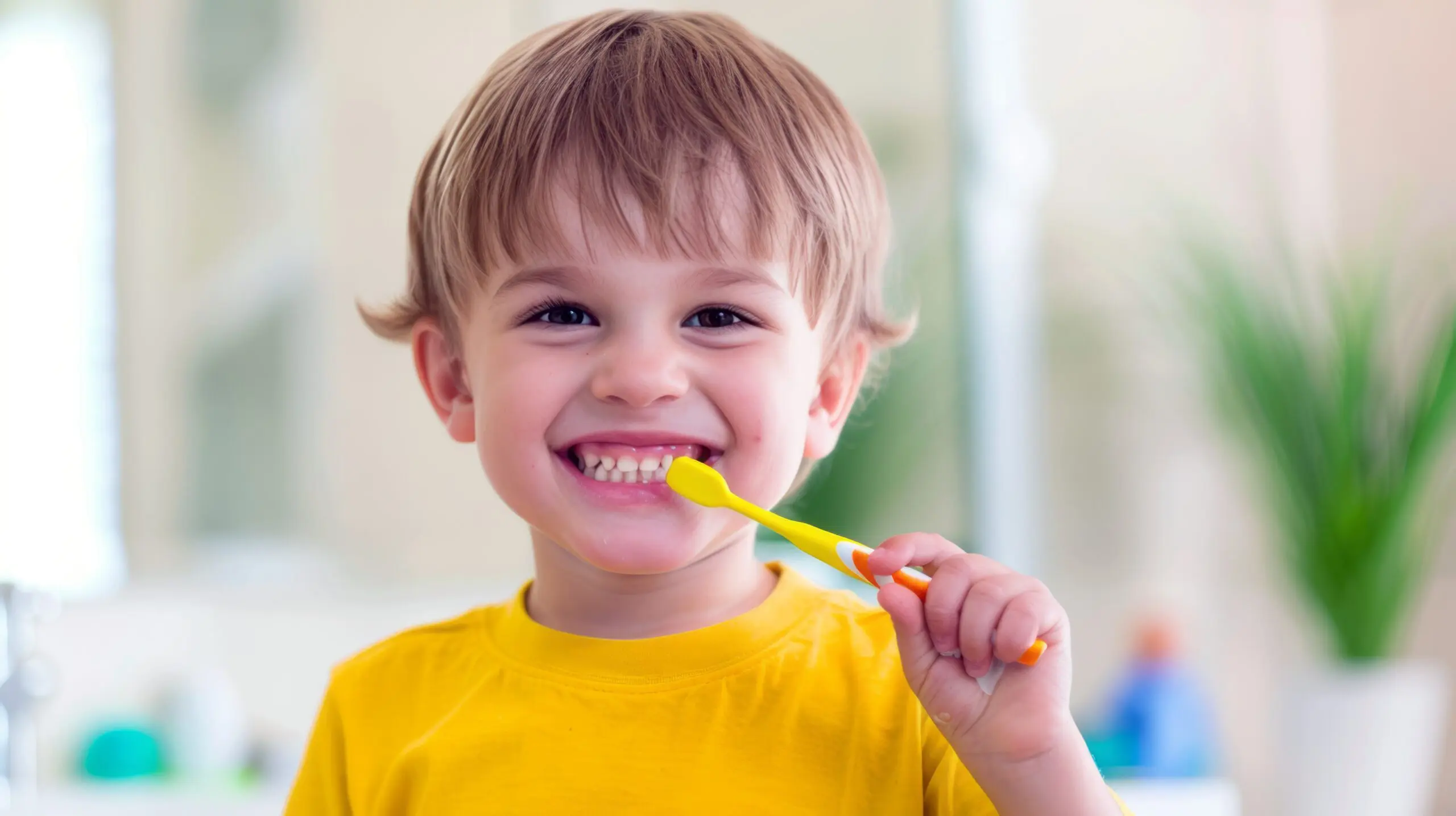 A smiling young child in a yellow shirt brushes their teeth in a brightly lit room, highlighting good oral health and restorative dentistry at Lake Sammamish Kids Dentistry - Pediatric Dentist in Sammamish, WA.