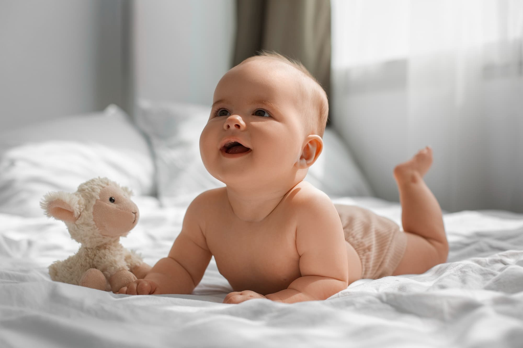 A baby smiles while lying on a bed next to a stuffed lamb, bathed in natural light—a heartwarming moment to cherish before their first visit to Lake Sammamish Kids Dentistry - Pediatric Dentist in Sammamish, WA by age 1.
