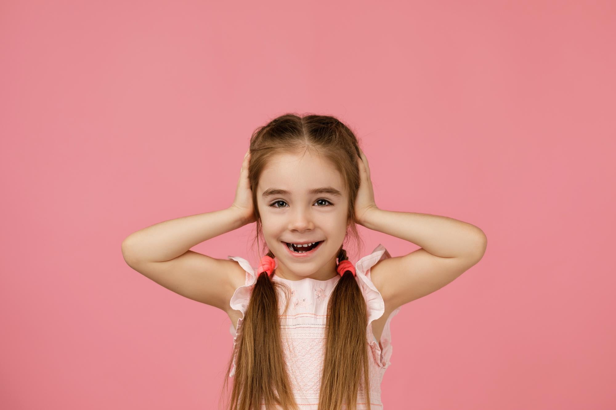 A young girl with brown hair in pigtails and a light pink dress smiles with her hands on her ears before a solid pink background, reflecting the happiness kids experience after visiting Lake Sammamish Kids Dentistry - Pediatric Dentist for preventive pediatric dental care in Sammamish, WA.