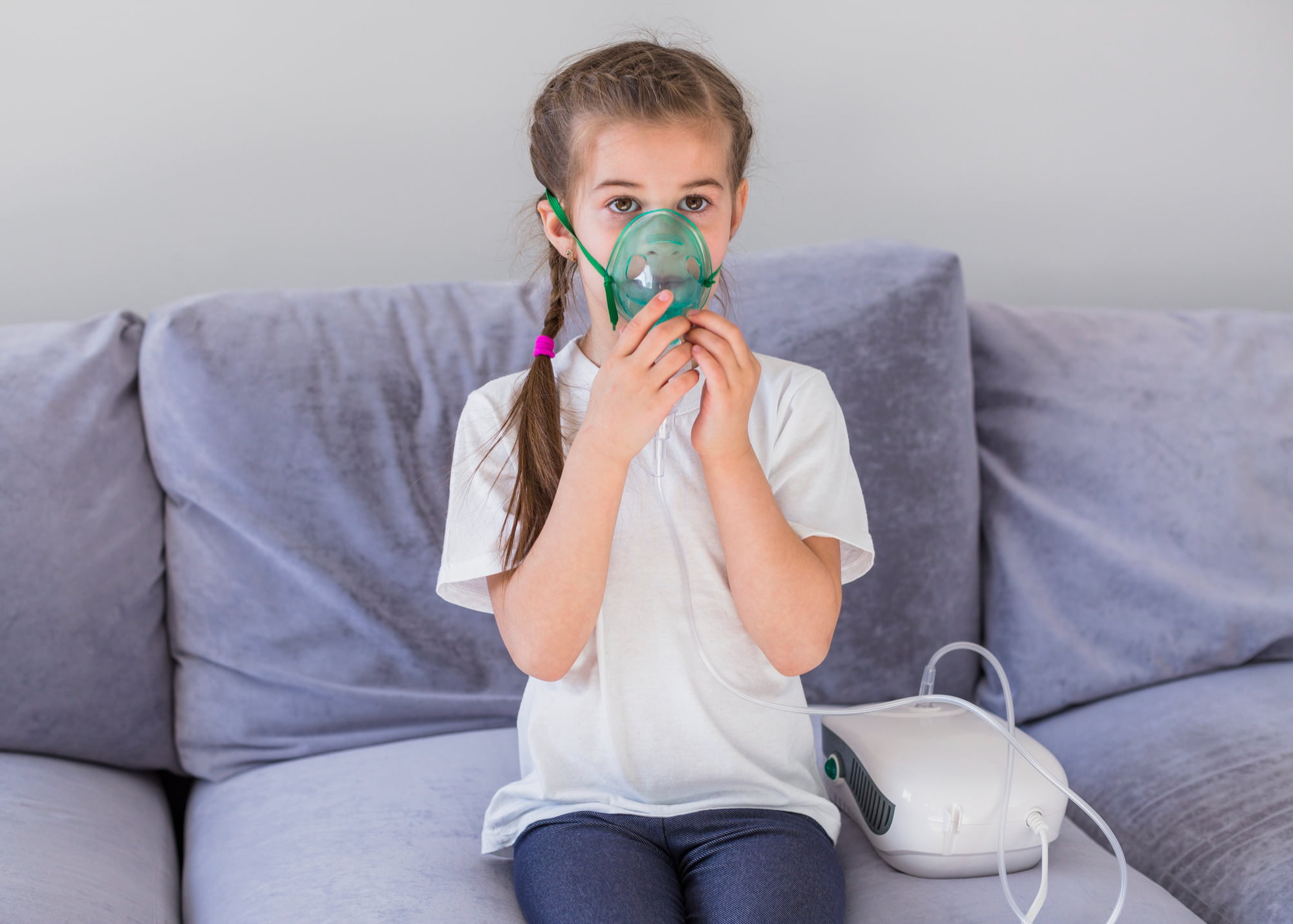 A young girl in a white shirt sits on a sofa using a nebulizer mask, resembling the nitrous oxide (laughing gas) equipment for kids at Lake Sammamish Kids Dentistry - Pediatric Dentist in Sammamish, WA.