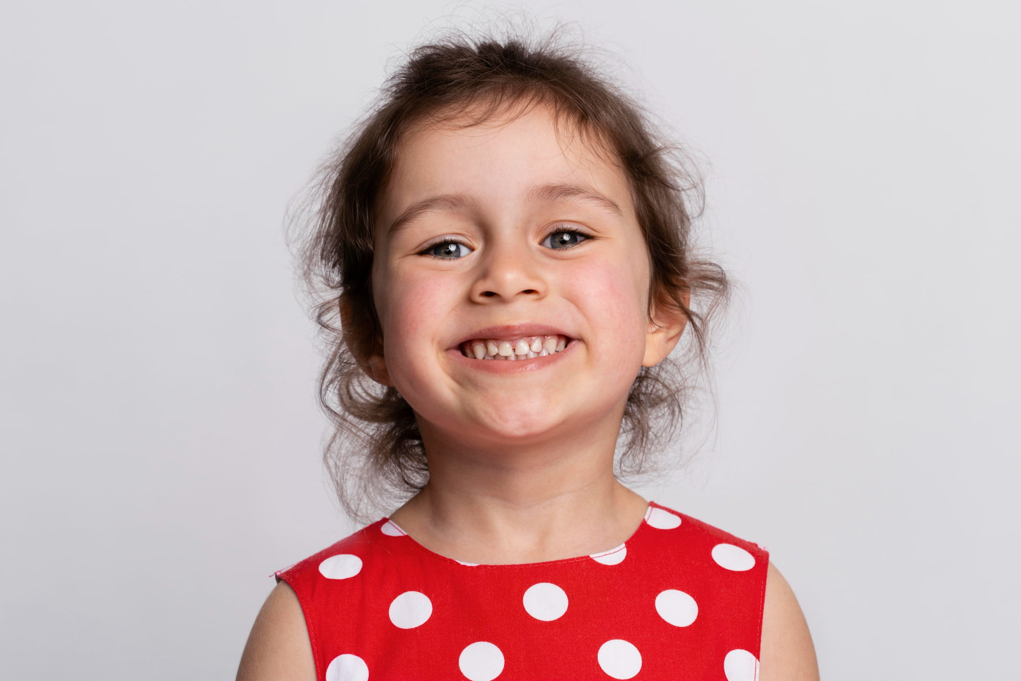 A smiling young child with brown hair in a red polka dot dress, set against a light background, illustrates how early orthodontics at Lake Sammamish Kids Dentistry - Pediatric Dentist in Sammamish, WA can benefit your child's smile through interceptive treatment.