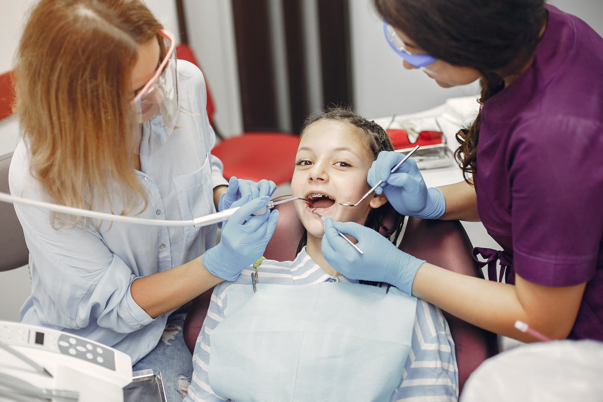 At Lake Sammamish Kids Dentistry - Pediatric Dentist in Sammamish WA, two dentists specializing in emergency pediatric care gently examine a young girl’s teeth as she sits in the dental chair wearing a bib, gloves, and safety glasses are used to ensure a safe and comfortable experience.