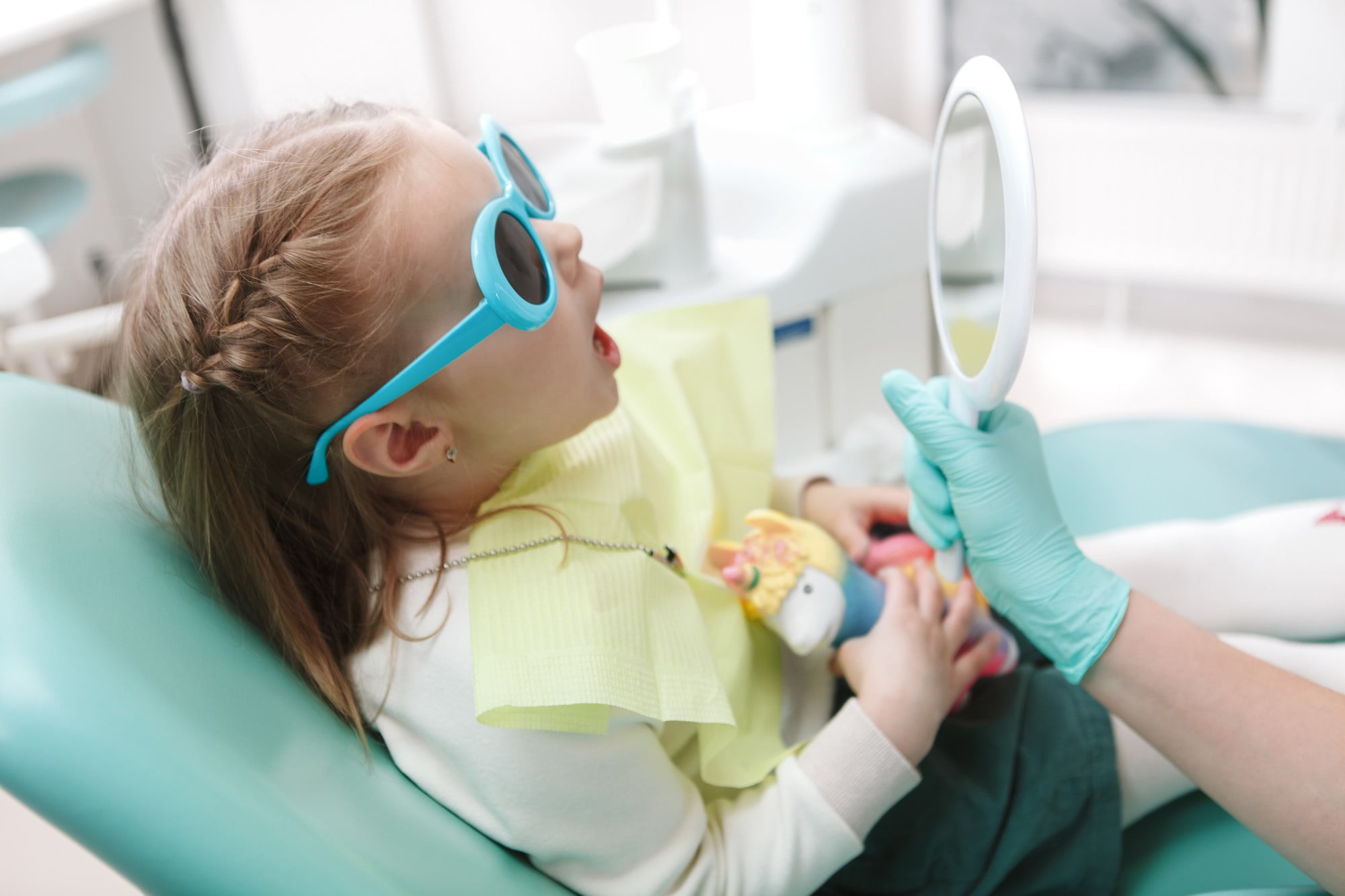 At Lake Sammamish Kids Dentistry - Pediatric Dentist in Sammamish WA, a young girl wearing blue sunglasses sits in the dental chair and examines her smile with a handheld mirror during her Soft-Tissue Magic: Frenectomies & Canker Sores appointment, guided by a gloved dental professional.