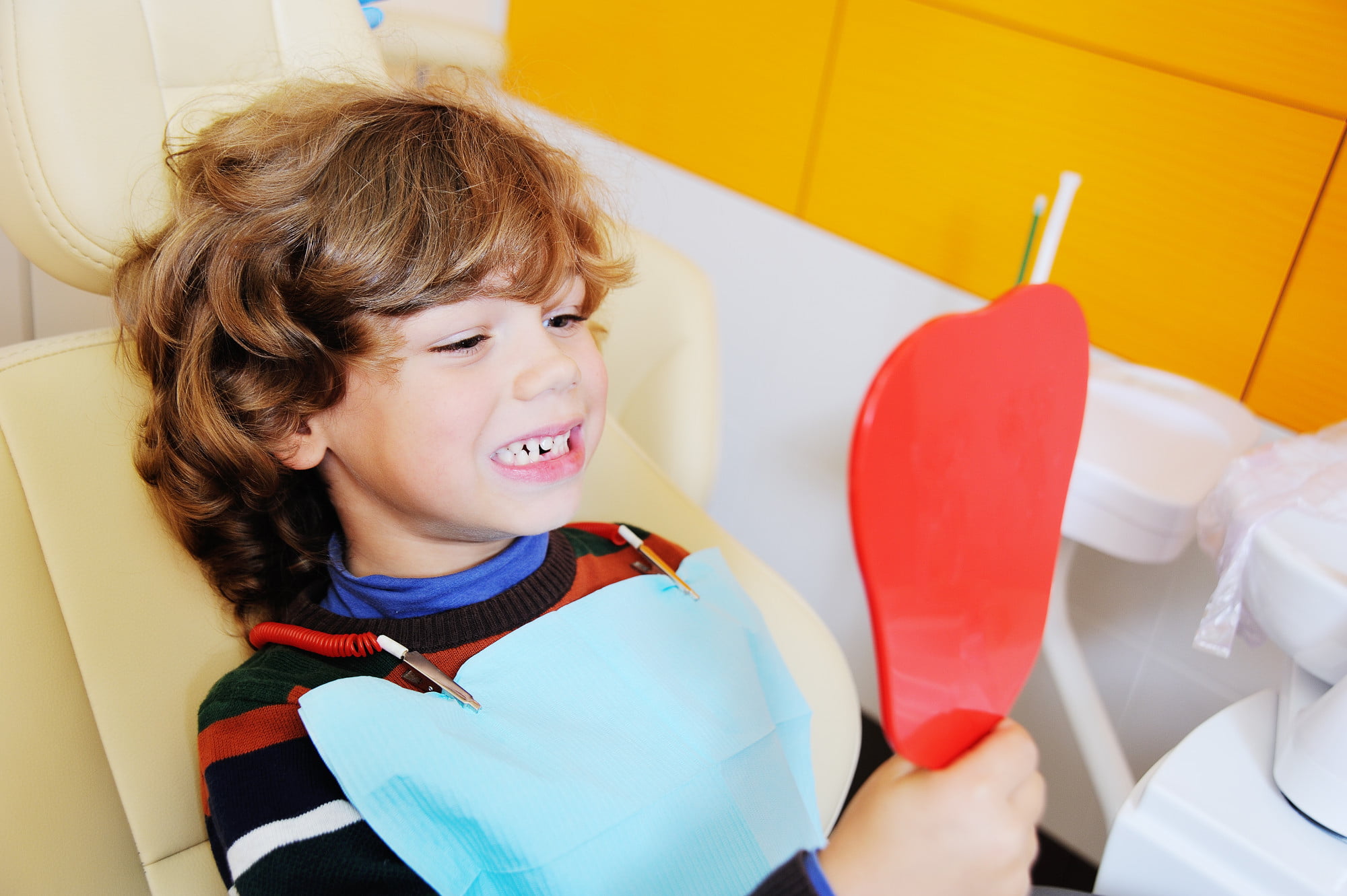 At Lake Sammamish Kids Dentistry - Pediatric Dentist in Sammamish WA, a young child sits in the dental chair wearing a striped sweater and bib, happily using a red heart-shaped mirror to check their teeth and identify potential tongue or lip ties.