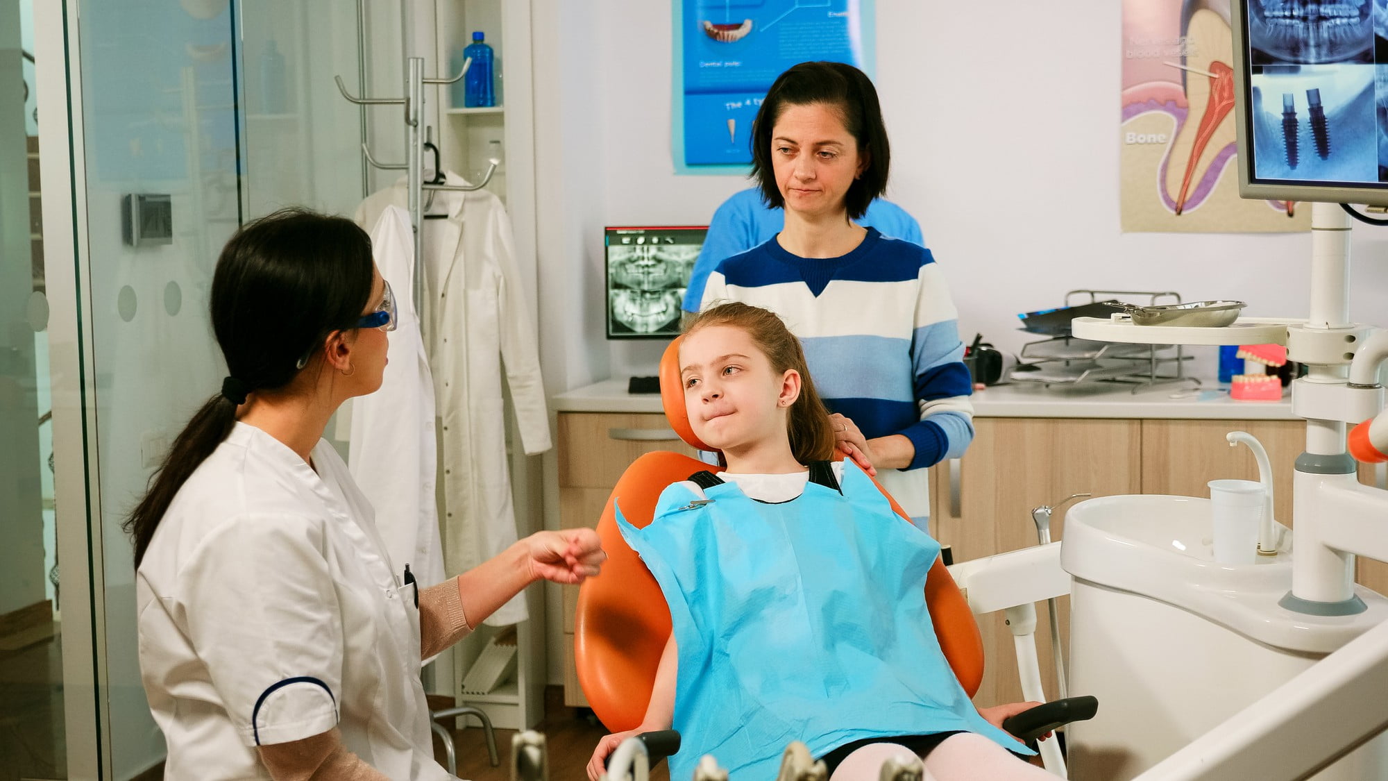 At Lake Sammamish Kids Dentistry - Pediatric Dentist in Sammamish WA, a young girl sits in a dental chair with a blue bib while the dentist explains the Preventive Pediatric Dental Care Checkup Schedule by age, accompanied by a woman standing beside her.