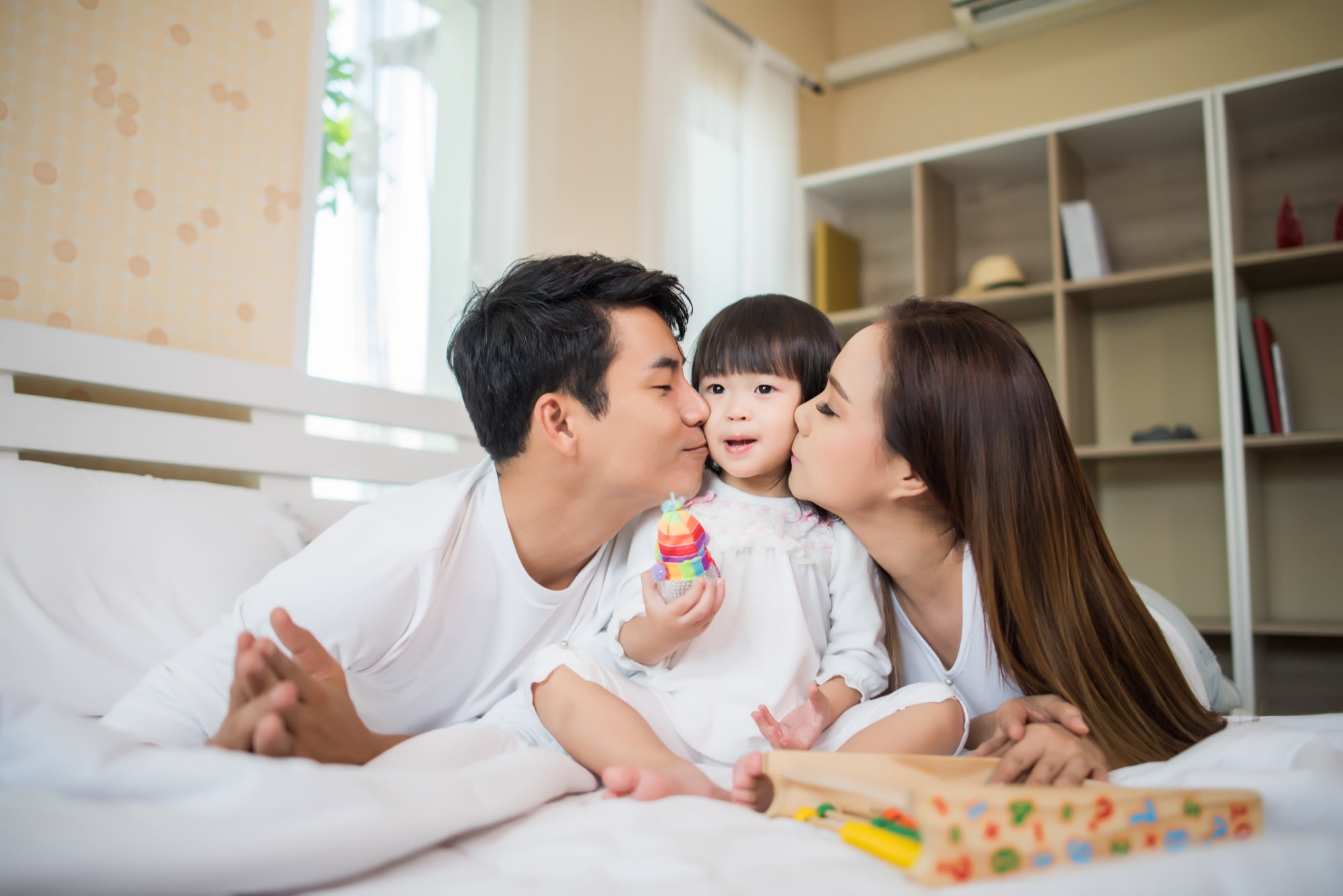 A child sits between two smiling adults on a bed at Lake Sammamish Kids Dentistry - Pediatric Dentist in Sammamish WA, receiving affectionate kisses on the cheeks. Holding a colorful toy, the child beams with happiness, while a wooden toy tray nearby highlights the warmth and sense of belonging that Apple Health Families: You Belong Here promotes.
