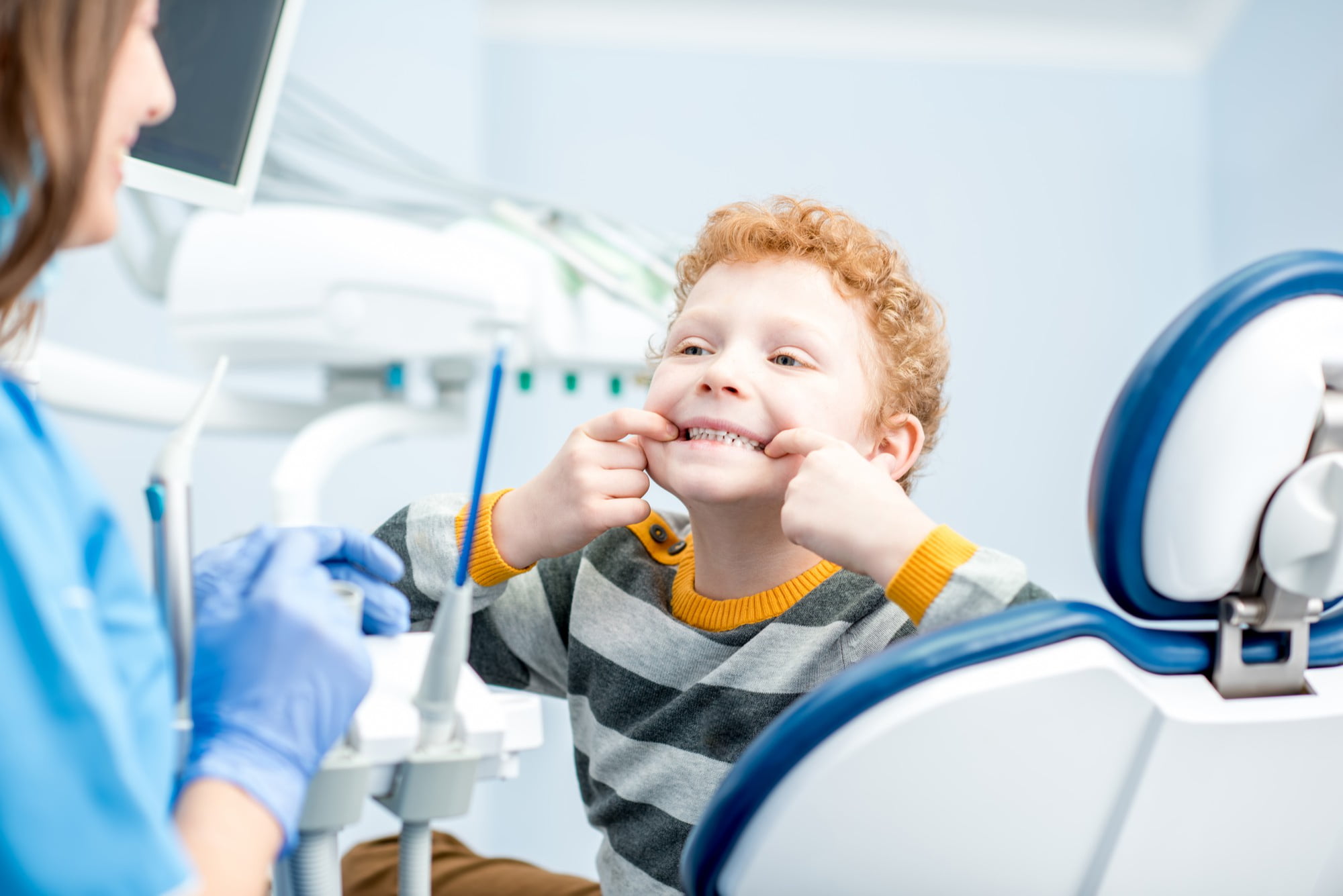 At Lake Sammamish Kids Dentistry - Pediatric Dentist in Sammamish WA, a young boy with curly red hair sits in the dental chair, pulling his mouth to show his teeth to a gloved emergency pediatric dentist—here's what to expect when you call.