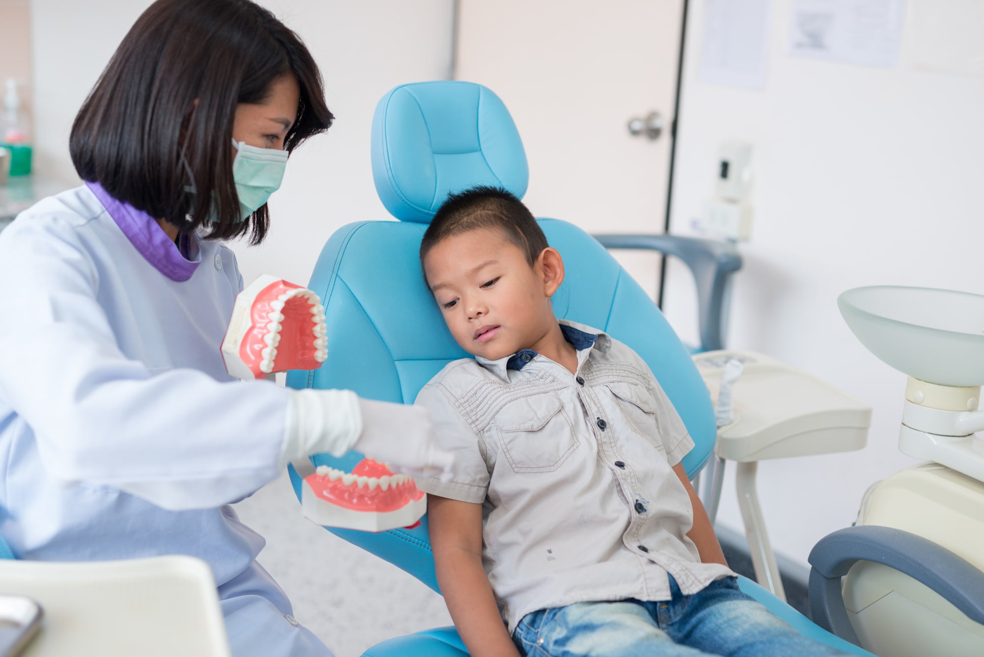 At Lake Sammamish Kids Dentistry - Pediatric Dentist in Sammamish WA, a masked dentist demonstrates proper oral care with a teeth model to a young boy sitting in a fully wheelchair-accessible dental chair.