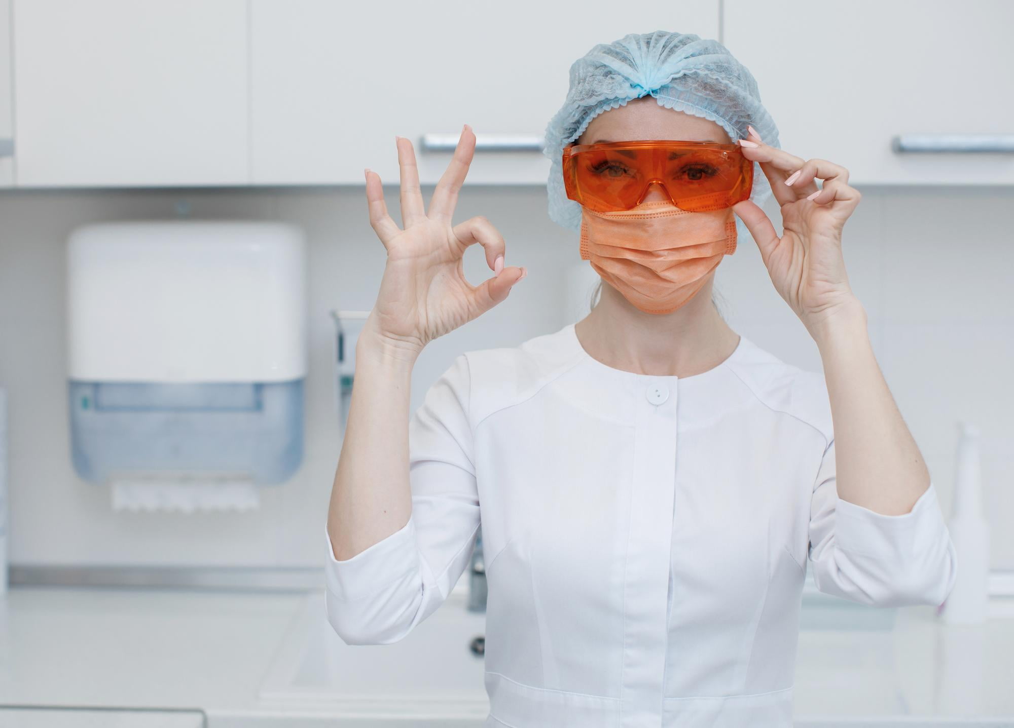 At Lake Sammamish Kids Dentistry - Pediatric Dentist in Sammamish WA, a dental professional—in a lab coat, hair cover, orange protective glasses, and face mask—makes an "OK" gesture in a clinical environment. PPO & Delta Dental Premier plans are accepted.