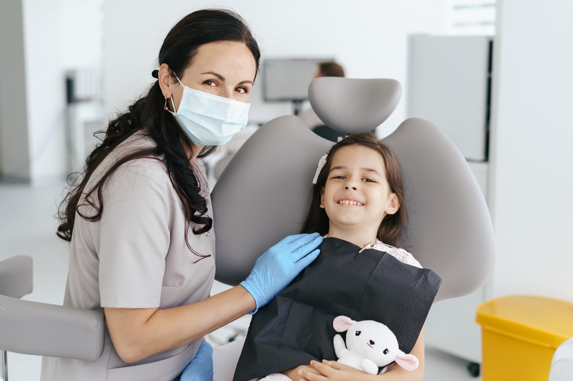 At Lake Sammamish Kids Dentistry - Pediatric Dentist in Sammamish WA, a masked and gloved dentist sits beside a smiling young girl holding a stuffed animal, emphasizing the importance of preventive pediatric dental care at your child’s first dental visit.
