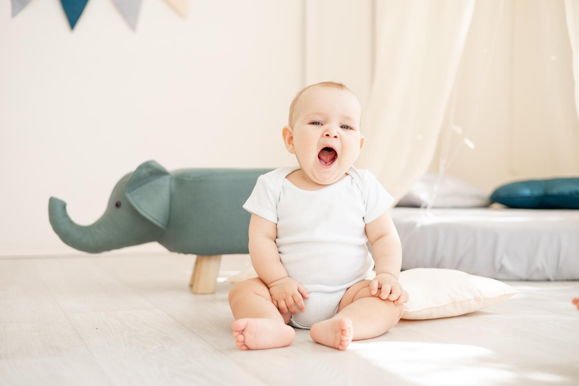 Reminding parents about oral hygiene for babies and toddlers, Lake Sammamish Kids Dentistry - Pediatric Dentist in Sammamish, WA shares an image of a yawning baby in a white onesie with a pillow and toy elephant nearby.