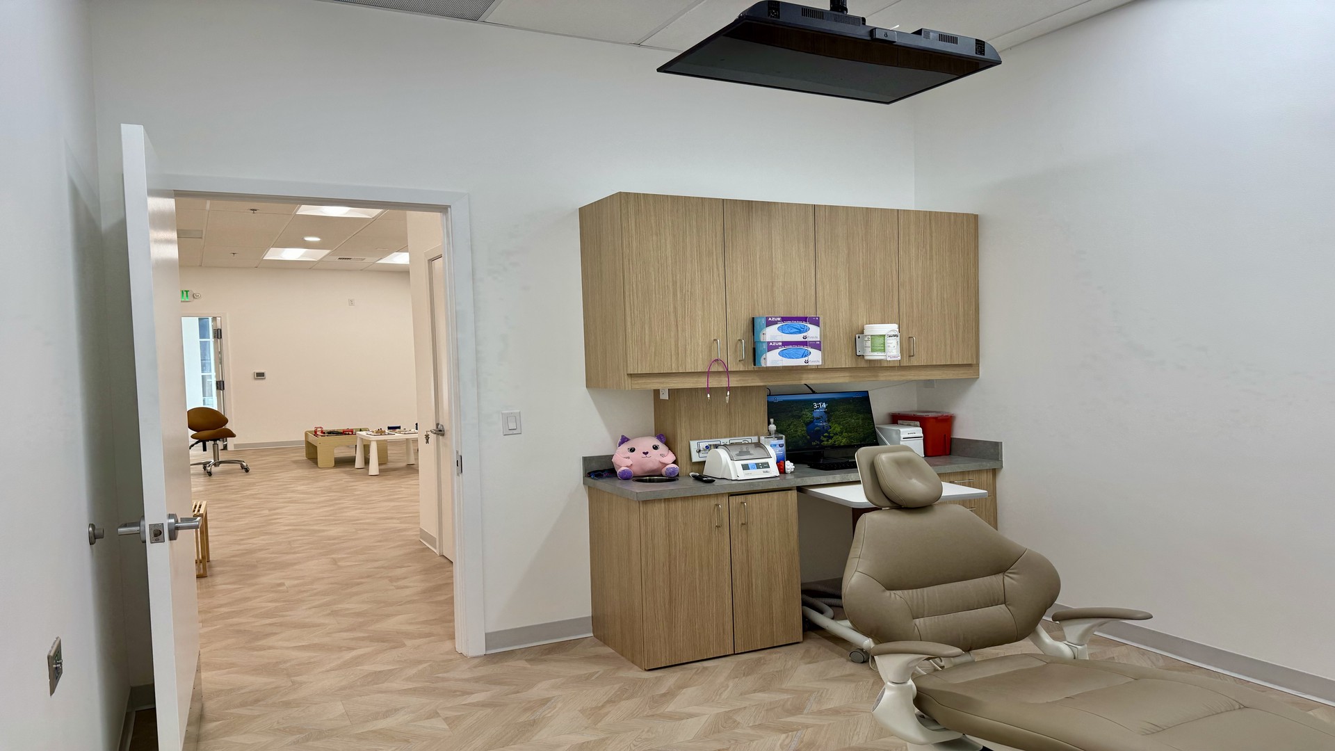 At Lake Sammamish Kids Dentistry - Pediatric Dentist in Sammamish, WA, the dental exam room features a modern dental chair, computer, and cabinets; through an open door, the waiting area includes seating and children's toys.