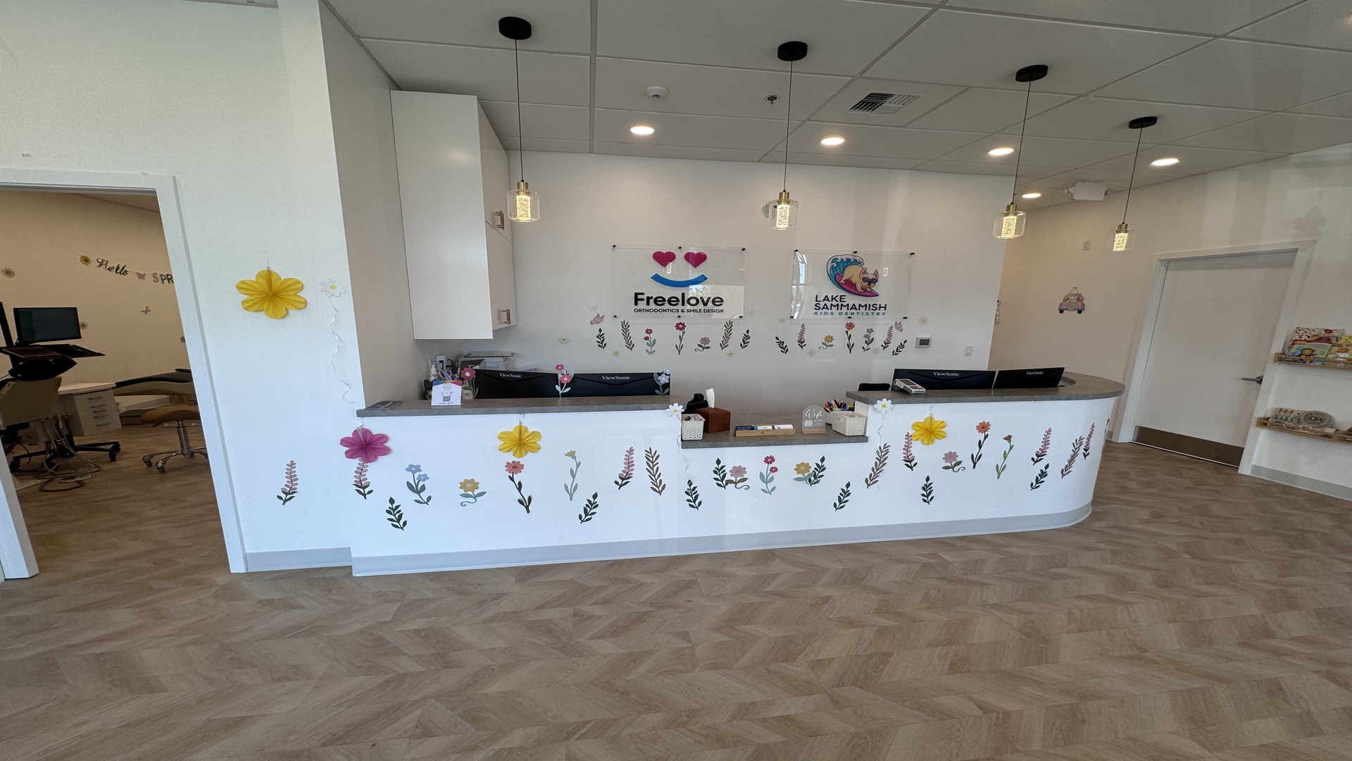 At Lake Sammamish Kids Dentistry - Pediatric Dentist in Sammamish, WA, the reception area features a curved white desk, floral wall decals, pendant lights, and signage for Freelove and Lacey Kishish, DDS. Through an open doorway, you can see an office and dental chair.