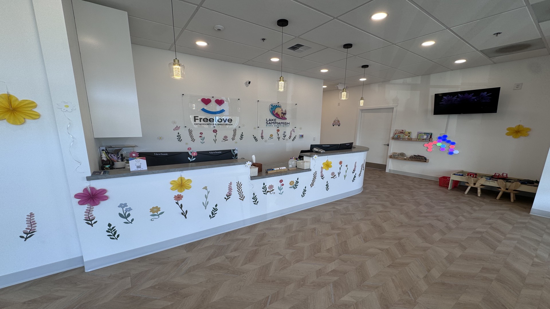 At Lake Sammamish Kids Dentistry - Pediatric Dentist in Sammamish WA, the reception area features a white counter adorned with colorful flower decals, pendant lights overhead, a kids’ play area to the right, and informational signs behind the desk—including a reminder: Emergency Care? Always Covered.