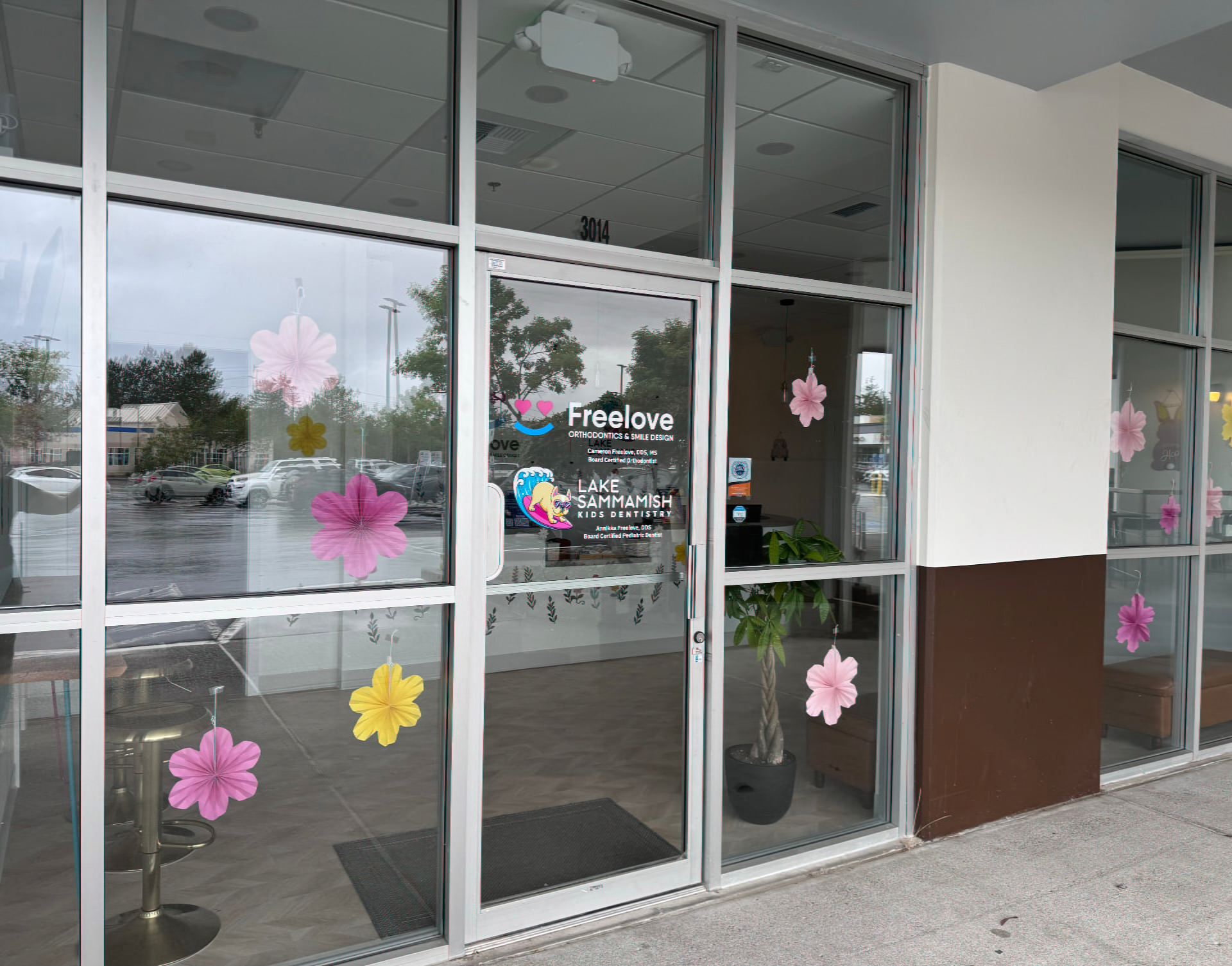 The glass storefront of Lake Sammamish Kids Dentistry - Pediatric Dentist in Sammamish, WA features colorful flower decals and entrance signs for Freelo Love and Lake Sammamish Family Dentistry, with a plant visible inside.