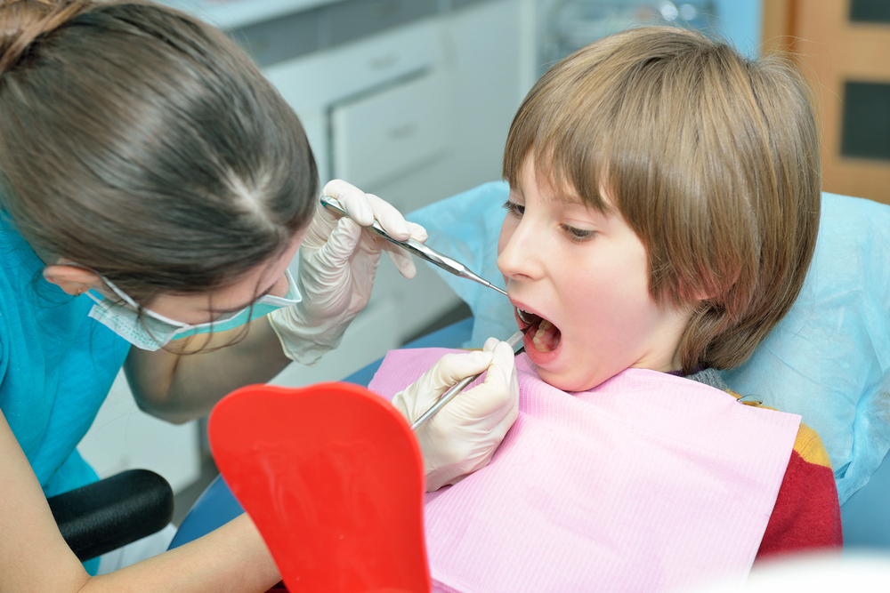 At Lake Sammamish Kids Dentistry - Pediatric Dentist in Sammamish, WA, a dentist uses dental tools to examine a young boy's teeth as he sits with a pink bib and holds a red mirror as one of the procedures of Pediatric Dental Emergencies in Sammamish, WA.