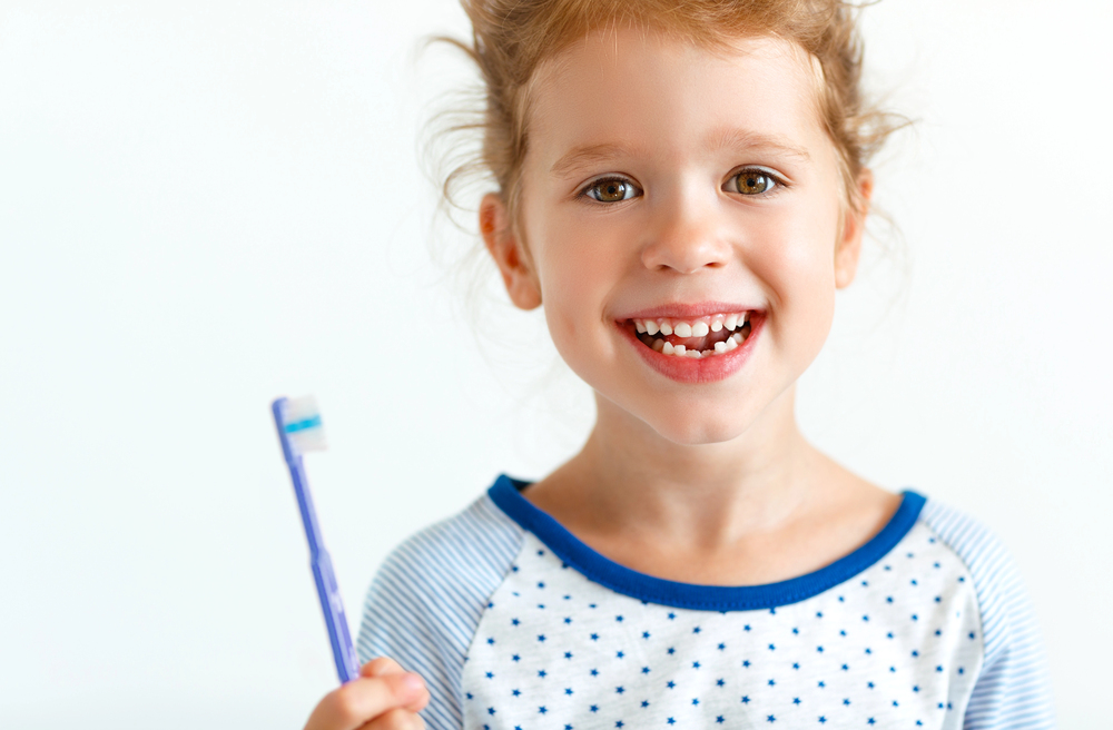 A young child, smiling with missing front teeth and holding a toothbrush, wears a blue and white polka-dotted shirt against a plain background—capturing Fluoride Safety for Children at Lake Sammamish Kids Dentistry - Pediatric Dentist in Sammamish, WA.
