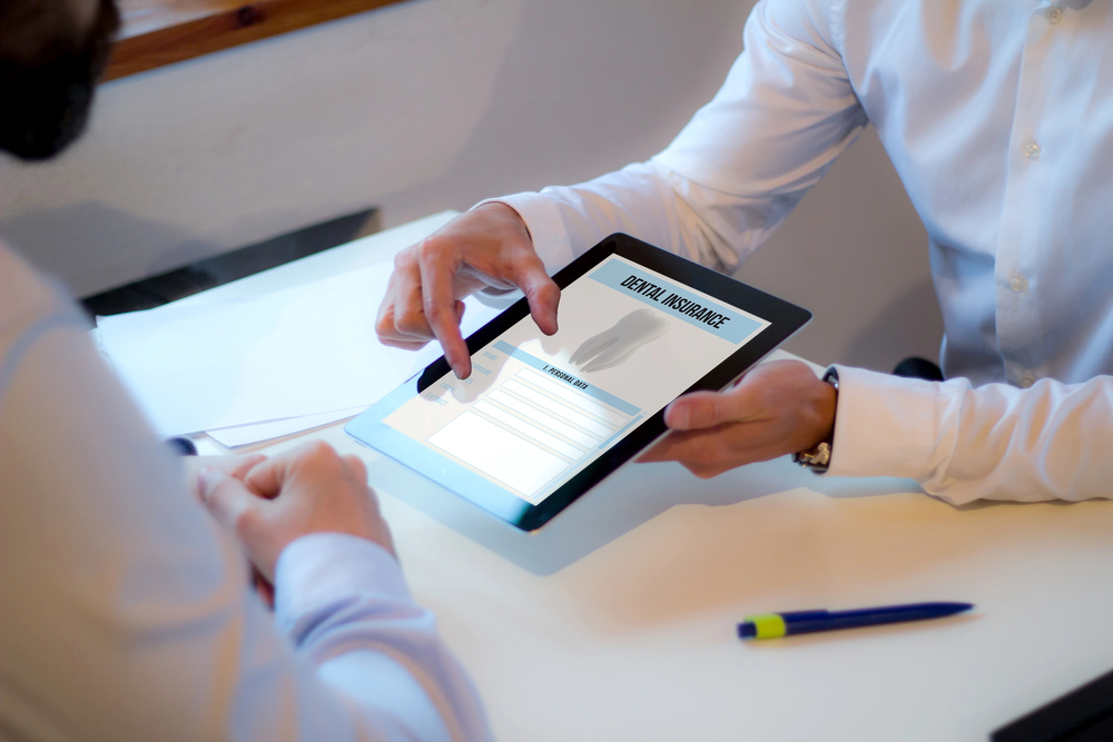 At a desk with papers and a pen, two people in business attire review an Apple Health Dental Coverage for Kids in Sammamish, WA form on a tablet at Lake Sammamish Kids Dentistry - Pediatric Dentist.