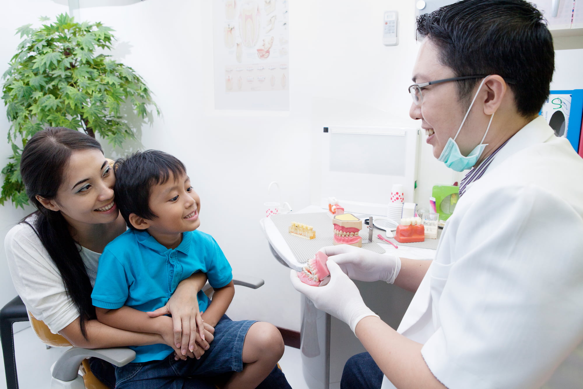 At Lake Sammamish Kids Dentistry - Pediatric Dentist in Sammamish WA, a dentist in a white coat uses Social Stories and visual supports to explain dental care with a model to a smiling young boy on his mother's lap in a welcoming office.