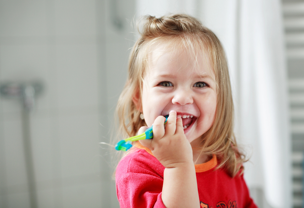 At Lake Sammamish Kids Dentistry - Pediatric Dentist in Sammamish, WA, a young child in a red shirt smiles while brushing their teeth with a toothbrush fluoride regardless The Fluoride Debate: A Pediatric Dentist’s Take in the bathroom.