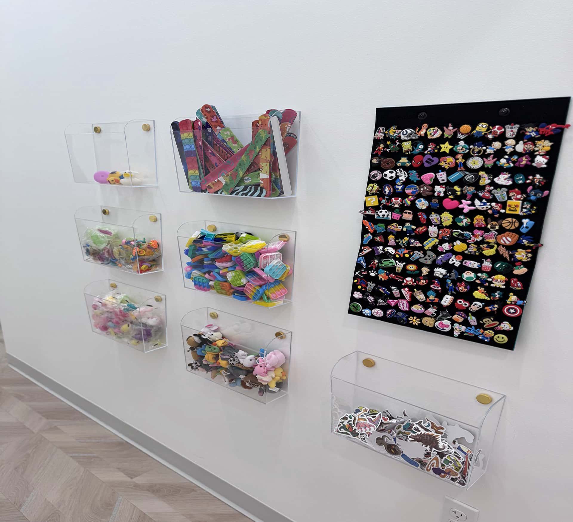 At Lake Sammamish Kids Dentistry - Pediatric Dentist in Sammamish, WA, clear wall-mounted organizers hold small toys and trinkets, while a black board showcases a vibrant collection of character-themed enamel pins — creating a cheerful space for young patients visiting an Infant & Baby Dentist.