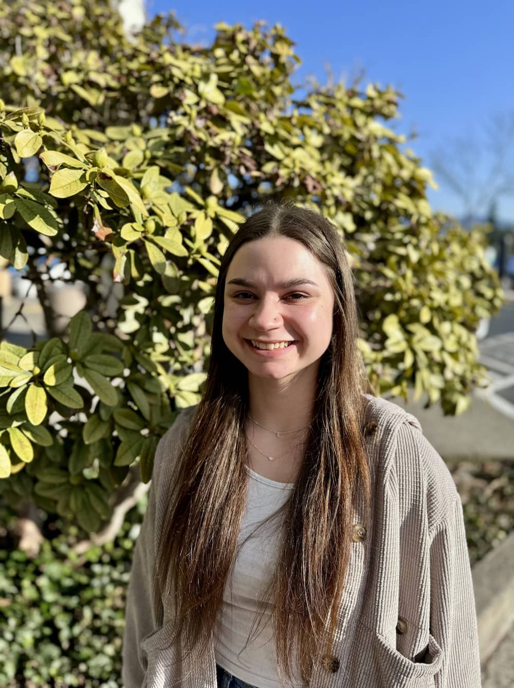 A young woman with long brown hair smiles warmly at the camera in front of a leafy green bush on a sunny day, reflecting the friendly atmosphere of Lake Sammamish Kids Dentistry - Pediatric Dentist in Sammamish, WA.