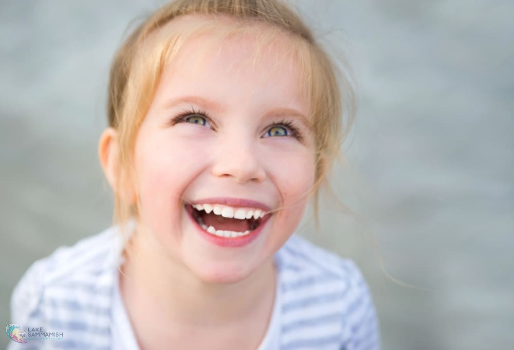A blonde girl smiles up, showing Fluoride Safety for Children on her teeth; at Lake Sammamish Kids Dentistry in Sammamish, WA, with a softly blurred background.