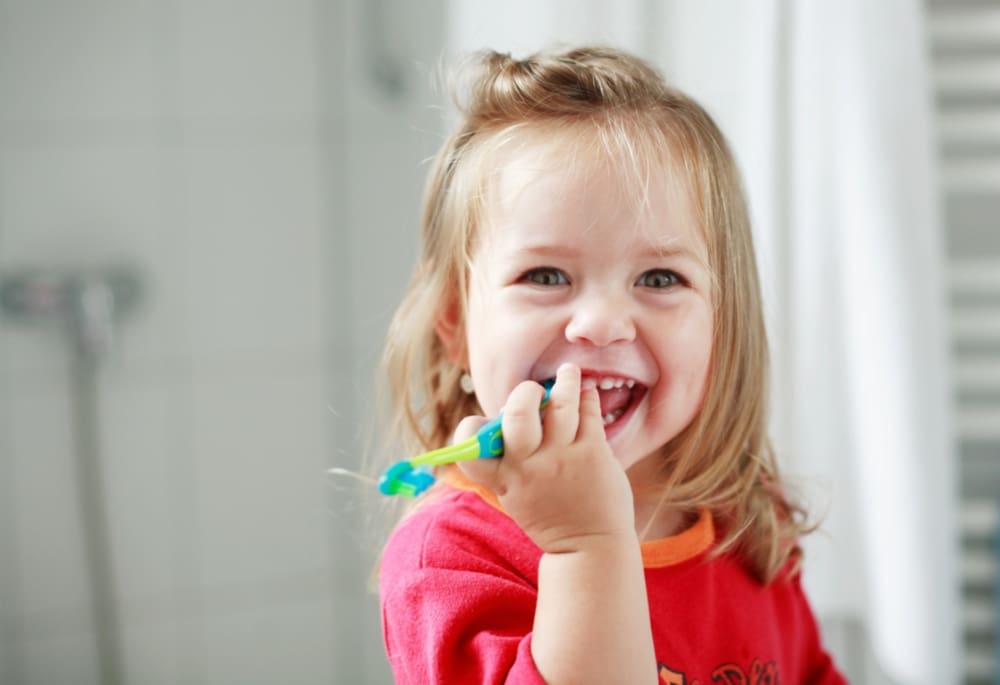 A young child in a red shirt smiles and chews on a toothbrush in a bright bathroom at Lake Sammamish Kids Dentistry in Sammamish, WA.