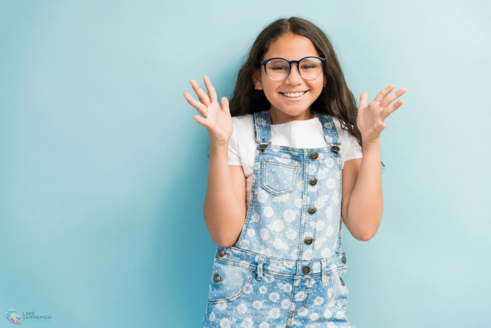 A smiling girl with long hair, glasses, and denim overalls raises her hands; Lake Sammamish Kids Dentistry in Sammamish, WA - Best Toothbrush for Kids by Age – Sammamish Parent Guide