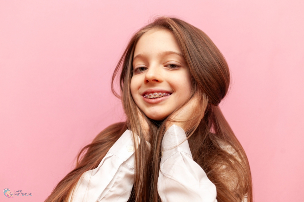 A smiling girl with long brown hair and braces stands against a pink background at Lake Sammamish Kids Dentistry in Sammamish, WA - Braces for Teens in Sammamish: What Parents Should Know